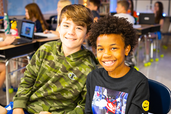 Two boys smiling and sitting at a desk in a classroom.
